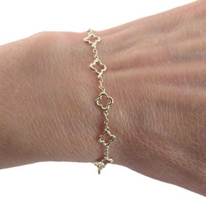 May include: A gold-colored bracelet featuring a series of openwork clover-shaped charms. The bracelet is delicate and appears to be made of a shiny metal, possibly gold. The bracelet is worn on a person's wrist.