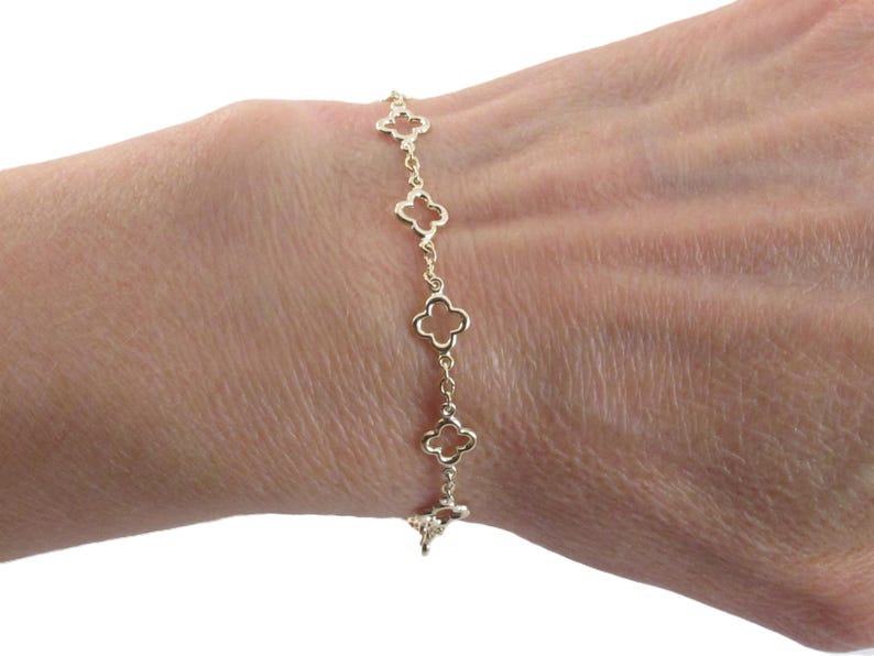 May include: A gold-colored bracelet featuring a series of openwork clover-shaped charms. The bracelet is delicate and appears to be made of a shiny metal, possibly gold. The bracelet is worn on a person's wrist.