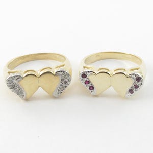 May include: Two gold rings, each featuring a double heart design. One ring has clear stones, the other has red stones. The rings have a gold band and a silver-colored accent around the hearts.