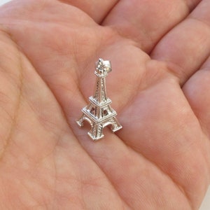 K&ouml;nnte beinhalten: Ein kleiner, silberner Eiffelturm-Anh&auml;nger. Der Anh&auml;nger ist eine detaillierte Miniatur-Nachbildung des ber&uuml;hmten Pariser Wahrzeichens mit aufwendigem Gitterwerk und einer spitzen Spitze. Der Anh&auml;nger wird in einer Hand gehalten, um seine Gr&ouml;&szlig;e und sein Design zu zeigen.