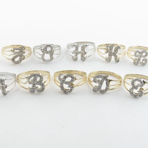 Könnte beinhalten: Eine Sammlung von zehn Initialenringen, jeder mit einem anderen Buchstaben des Alphabets. Die Ringe sind in Gold- und Silbertönen erhältlich, wobei die Buchstaben mit kleinen, funkelnden Steinen besetzt sind. Die Ringe werden auf einer weißen Oberfläche präsentiert.