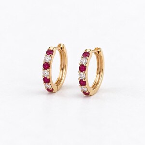 May include: A pair of gold hoop earrings featuring an alternating pattern of round, deep red rubies and clear, sparkling white diamonds set along the front curve of each hoop, secured with a hinged closure mechanism.