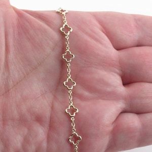 May include: A gold-colored chain bracelet featuring a series of openwork clover-shaped charms. The bracelet is displayed against a skin-toned background, showcasing the delicate design and the warm hue of the metal.
