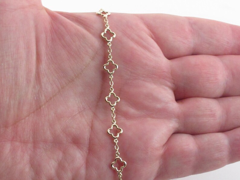 May include: A gold-colored chain bracelet featuring a series of openwork clover-shaped charms. The bracelet is displayed against a skin-toned background, showcasing the delicate design and the warm hue of the metal.
