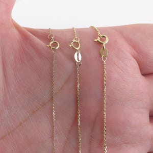 May include: Three gold chains of different lengths, shown on a hand. The chains are all the same style, with a delicate, linked design.