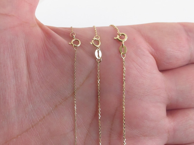 May include: Three gold chains of different lengths, shown on a hand. The chains are all the same style, with a delicate, linked design.