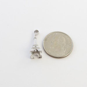 K&ouml;nnte beinhalten: Ein silberner Eiffelturm-Anh&auml;nger neben einer US-amerikanischen Vierteldollarm&uuml;nze. Der Anh&auml;nger ist eine detaillierte Miniatur des ber&uuml;hmten Pariser Wahrzeichens. Die M&uuml;nze ist silberfarben.