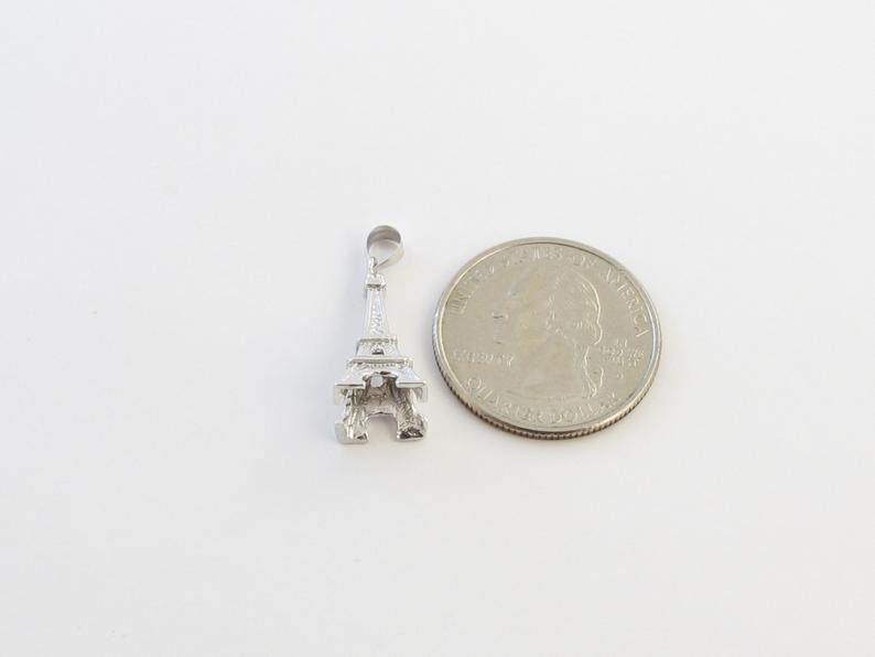 K&ouml;nnte beinhalten: Ein silberner Eiffelturm-Anh&auml;nger neben einer US-amerikanischen Vierteldollarm&uuml;nze. Der Anh&auml;nger ist eine detaillierte Miniatur des ber&uuml;hmten Pariser Wahrzeichens. Die M&uuml;nze ist silberfarben.