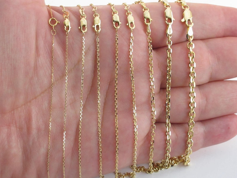 May include: A selection of gold necklaces with various chain styles, including cable, box, and rolo chains. Each necklace is displayed with its clasp open, showcasing the different designs and textures of the chains. The necklaces are held in a hand against a white background.