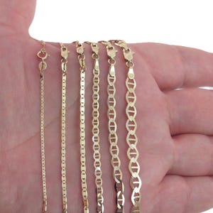 May include: A selection of gold-coloured chain necklaces in various styles and thicknesses. The chains are displayed against a light background. The chains are of different lengths and designs, including box, curb, and mariner styles.