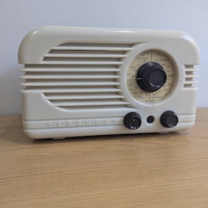 May include: A cream-coloured vintage Ferguson radio with a rounded top and horizontal vent design. The radio has a circular dial with black knobs and the brand name "FERGUSON" across the front. It sits on a wooden surface.