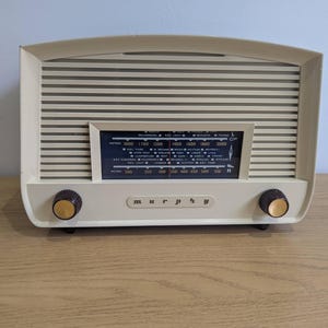 May include: A vintage cream-coloured radio with a horizontal striped speaker cover and a dark blue tuning dial. The radio has two brown knobs and the name "murphy" printed on the front. It sits on a wooden surface.