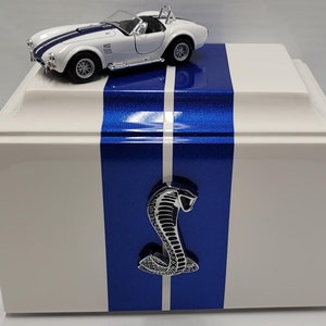 May include: White wooden cremation urn with blue racing stripes and a silver cobra emblem. A white toy car sits on top of the urn.