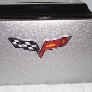 May include: A silver box with a black top, featuring the Chevrolet Corvette logo. The logo is a stylized emblem with a checkered flag and a red flag with a gold cross and fleur-de-lis.