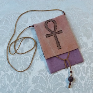 May include: A small, pink and purple fabric pouch with a brown Ankh symbol embroidered on the front. The pouch is attached to a brown cord with a dark brown bead.