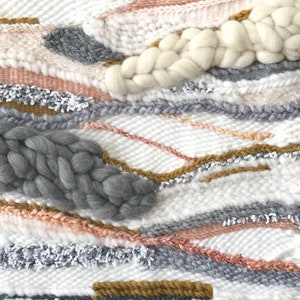 Close-up of a woven wall hanging featuring a textured, abstract design. The piece incorporates various shades of gray, pink, white, and gold threads, along with chunky wool elements in gray and off-white. The weaving technique creates a layered, dimensional effect with varying textures and colors.  The overall aesthetic is modern and minimalist, suitable for contemporary home decor.