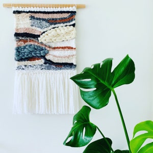 A woven wall hanging features abstract shapes in shades of gray, blush pink, copper, and white. The wall hanging has a white fringe at the bottom.  A monstera deliciosa plant is partially visible in the lower right corner.