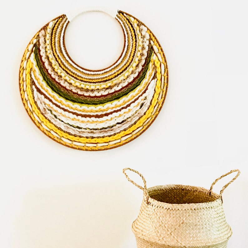 A large, circular wall hanging crafted from woven materials in shades of beige, yellow, and olive green. The piece features concentric bands of varying textures and colors, creating a visually appealing layered effect. Below it is a light beige seagrass basket with two handles.