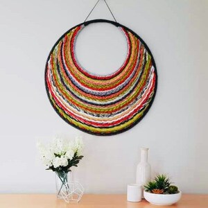 A circular wall hanging features multicolored yarn woven in concentric circles, creating a rainbow effect. The yarn colors include red, orange, yellow, green, gray, and white. The weaving is attached to a black metal circular frame. The wall hanging has a modern and bohemian aesthetic, suitable for home decor.