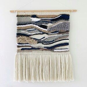A woven wall hanging features an abstract landscape design in shades of gray, beige, and navy blue. The piece is handcrafted with a textured, multi-colored yarn and has a long fringe at the bottom.  The wall hanging is rectangular and is displayed against a white background. It is mounted on a light brown wooden dowel.