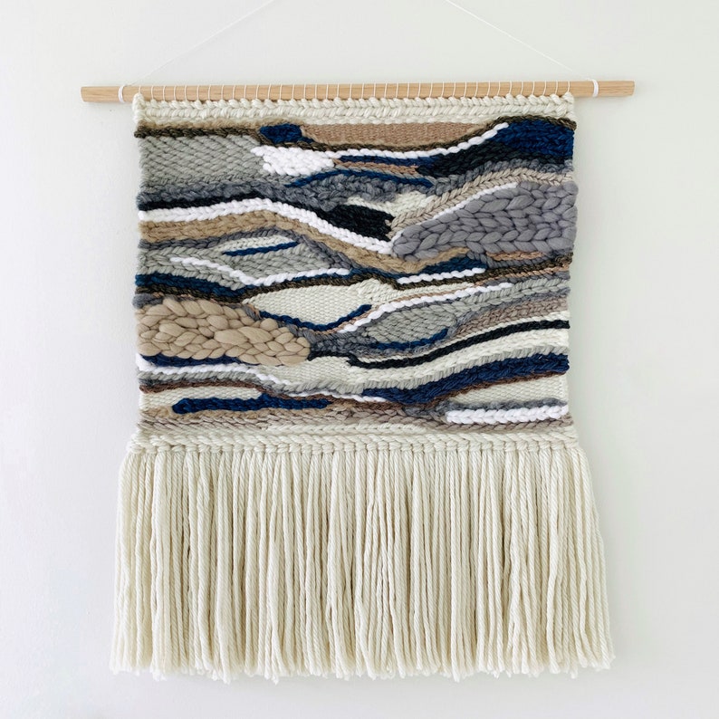 A woven wall hanging features an abstract landscape design in shades of gray, beige, and navy blue. The piece is handcrafted with a textured, multi-colored yarn and has a long fringe at the bottom.  The wall hanging is rectangular and is displayed against a white background. It is mounted on a light brown wooden dowel.