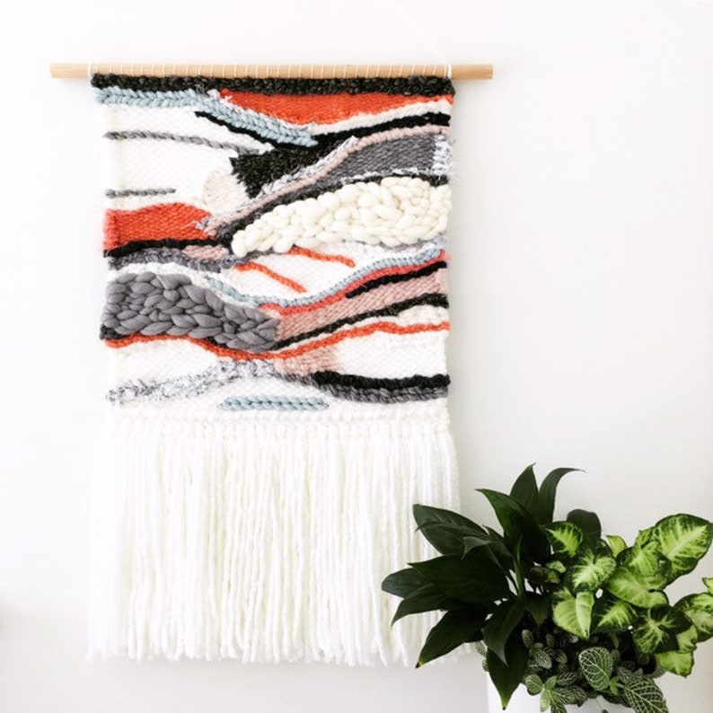 A woven wall hanging features an abstract landscape design in shades of orange, gray, white, and black. The piece is adorned with a long fringe at the bottom, adding texture and visual interest. The wall hanging is mounted on a simple wooden rod, suggesting a modern and minimalist aesthetic.  The colors are muted and earthy, creating a calming and sophisticated atmosphere. The texture is varied, with sections of tightly woven fabric and others with a more loose, open weave.