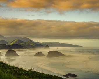 May include: A scenic coastal landscape featuring a beach, ocean, and large rock formations. The sky is filled with golden clouds, and the scene is bathed in soft, diffused light. Green hills and a shoreline are visible in the background.