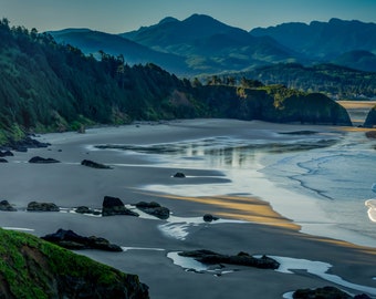 May include: A scenic coastal landscape featuring a sandy beach, ocean waves, and rocky formations. Lush green hills and cliffs border the shore, with a backdrop of distant mountains under a blue sky. The image captures a serene natural environment.