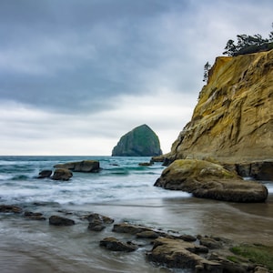 May include: A scenic coastal landscape featuring a rocky beach, ocean waves, and a large rock formation in the distance. A tall, layered cliff is on the right, and the sky is overcast.