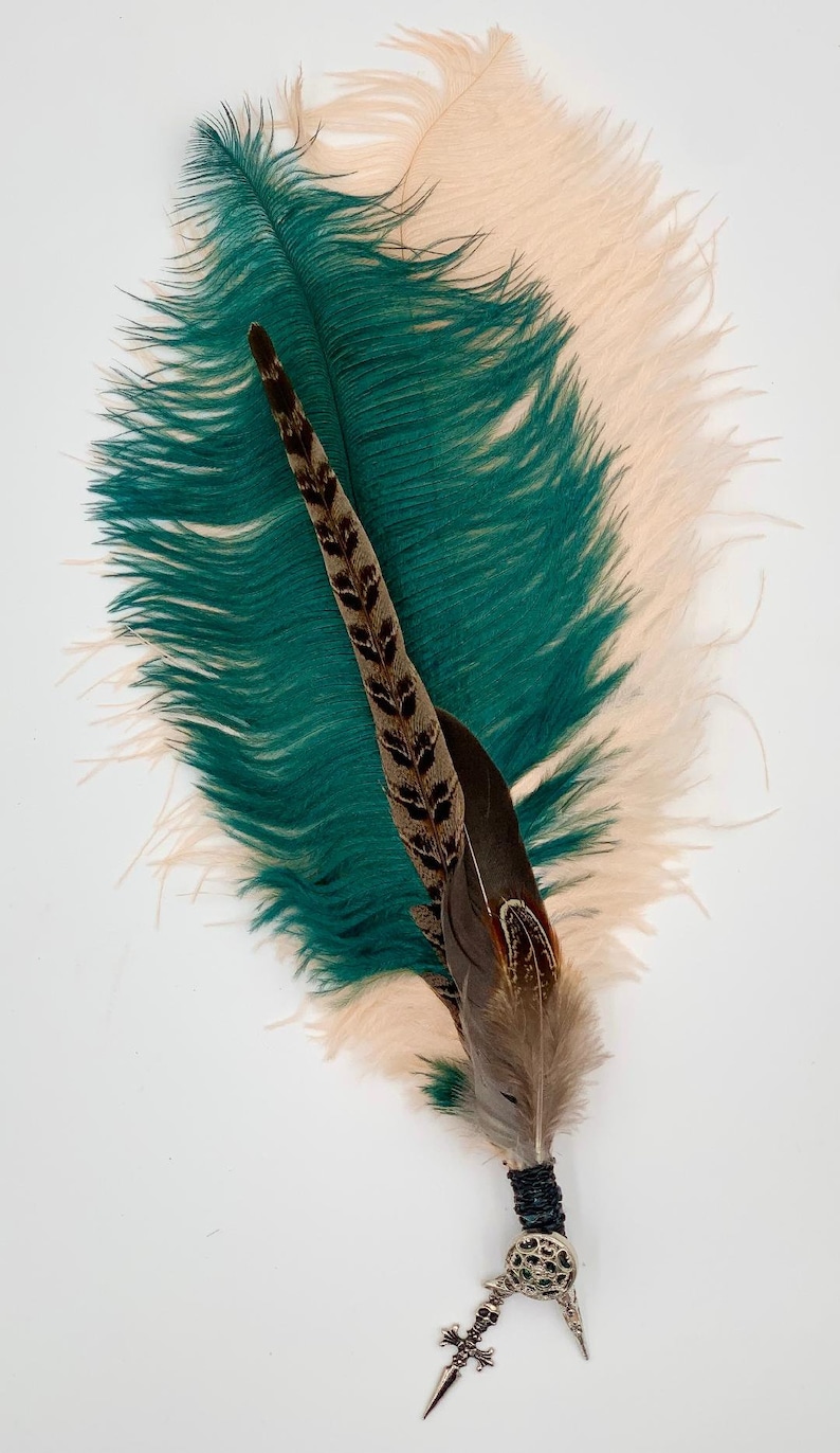 Op de afbeelding: Een groene en beige struisvogelveer met een bruine en zwarte fazantenveer bevestigd aan een zilveren en zwarte decoratieve pin.