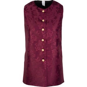 May include: A sleeveless vest in a deep burgundy color featuring a tonal floral jacquard pattern throughout. The garment has a round neckline and a front button closure with five circular gold-toned buttons. The interior lining is black.