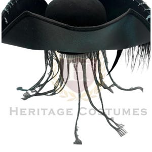 Puede incluir: Un sombrero de tres picos negro con un ala ancha y curva con costuras en color verde azulado a lo largo de los bordes. El sombrero est&aacute; adornado con tiras de tela negra largas y finas que cuelgan de la base de la copa y plumas negras.