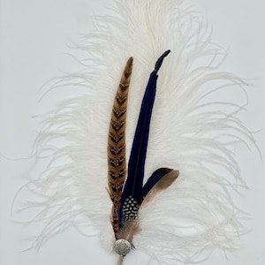 Op de afbeelding: Een witte struisvogelveer met een blauwe en bruine fazantenveer accent. De veren zijn bevestigd aan een zilveren pin met een decoratief cirkelvormig ontwerp.