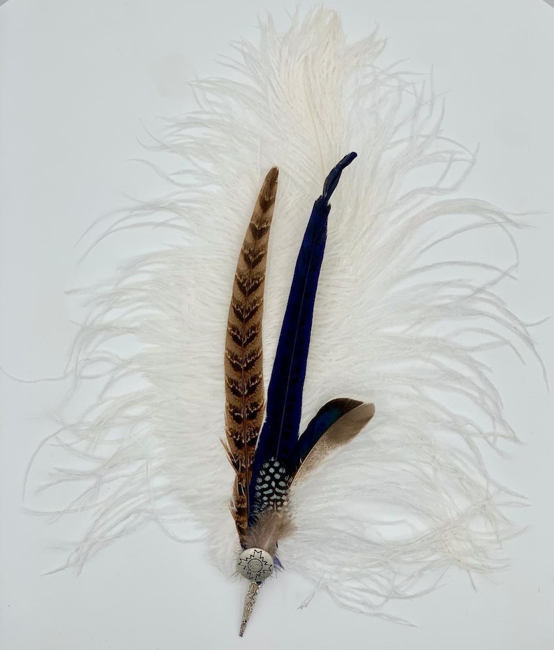 Op de afbeelding: Een witte struisvogelveer met een blauwe en bruine fazantenveer accent. De veren zijn bevestigd aan een zilveren pin met een decoratief cirkelvormig ontwerp.