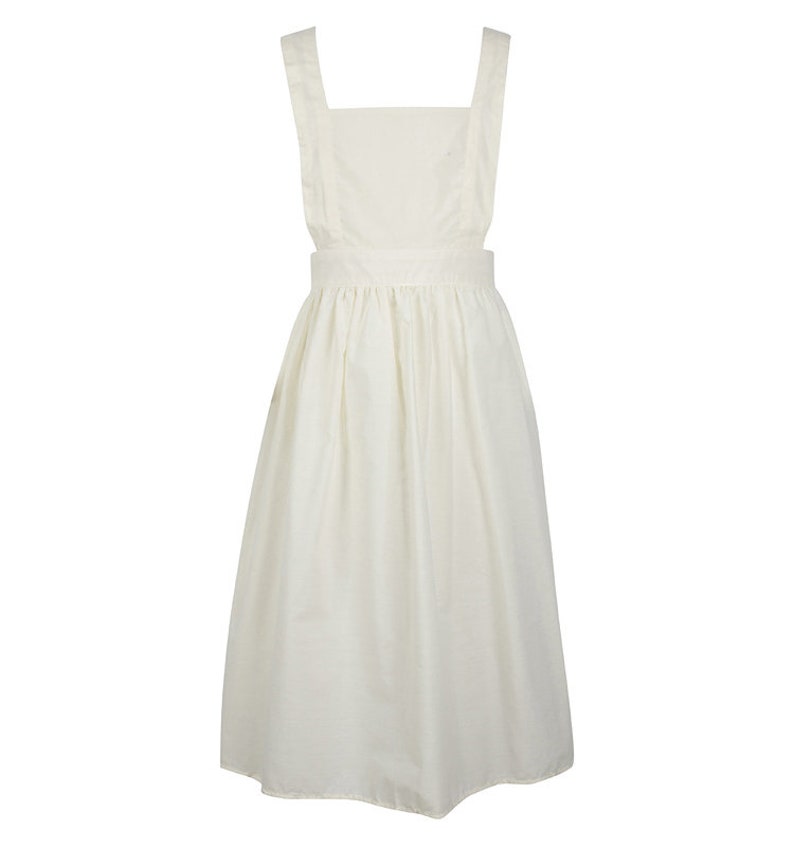 May include: A solid off-white pinafore dress featuring a square neckline, wide shoulder straps, and a fitted waistband that transitions into a gathered, knee-length skirt. The garment is constructed from a plain, light-coloured fabric with a simple design.