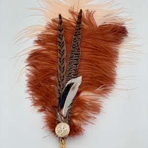Op de afbeelding: Een bruine, perzikkleurige en witte verenpluim met een gouden pin en een ronde, witte knoop met een decoratief ontwerp.