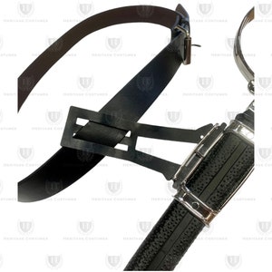 May include: A black leather belt with a silver-toned metal buckle and a black scabbard featuring a textured surface and silver-toned metal accents. The scabbard is attached to the belt by a black rectangular frame connector piece.