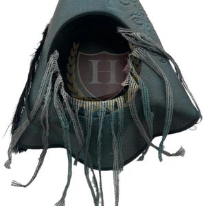 Puede incluir: Un sombrero de tres picos en color verde azulado oscuro con acabado desgastado, detalles de plumas negras y tiras de malla gris colgantes. El interior muestra un emblema circular con una letra H rodeada por una corona de laurel dorada.
