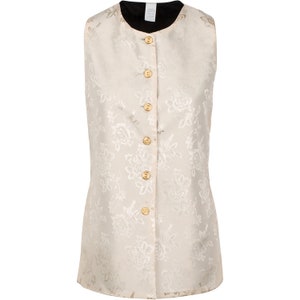 May include: A sleeveless vest in a cream-colored fabric featuring a tonal floral jacquard pattern. The garment has a round neckline, a black interior lining, and a front closure secured by six round, gold-toned buttons with a textured surface.