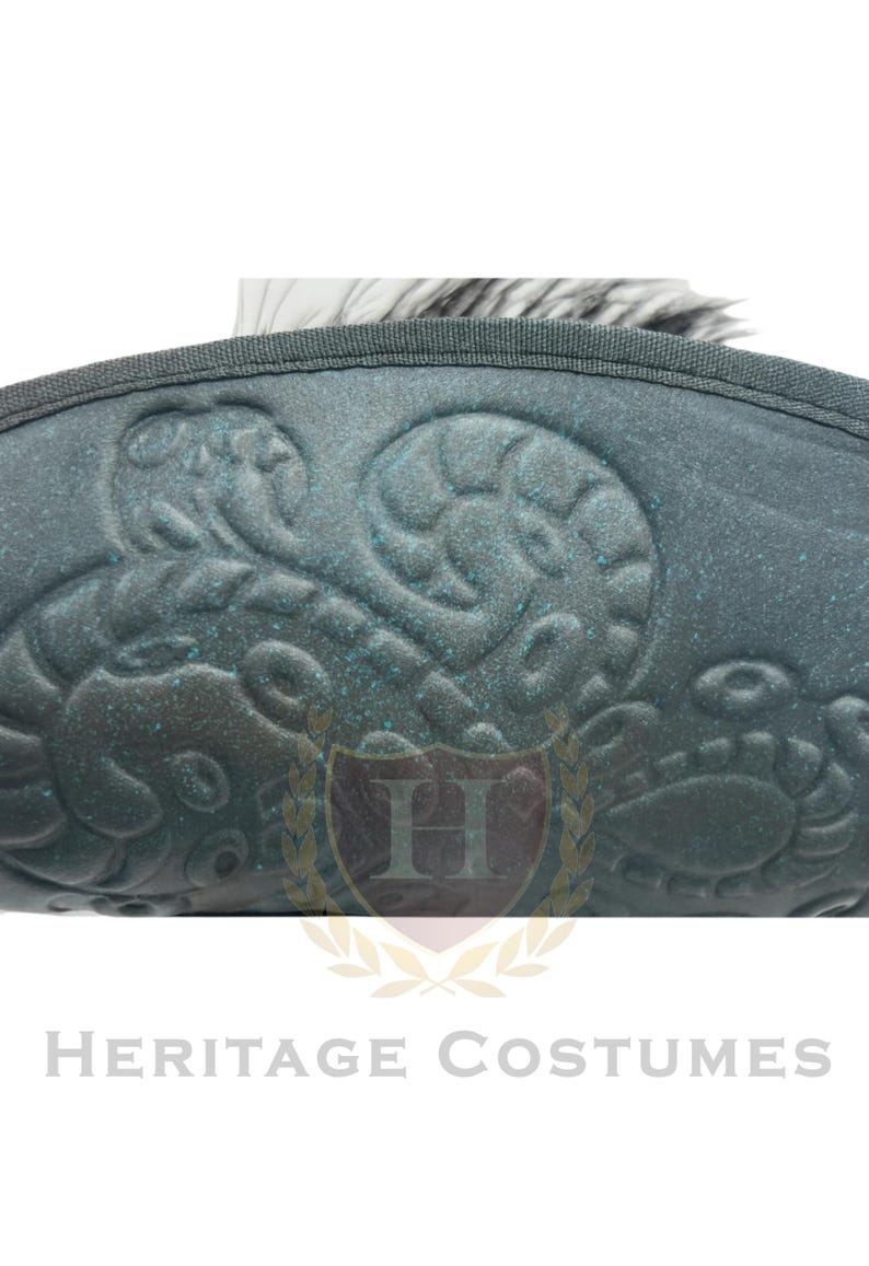 Puede incluir: Un sombrero de color verde azulado oscuro con un patr&oacute;n de remolinos en relieve en la superficie. El sombrero tiene un borde cosido y una pluma blanca que sobresale por la parte superior. La superficie tiene un brillo met&aacute;lico sutil.