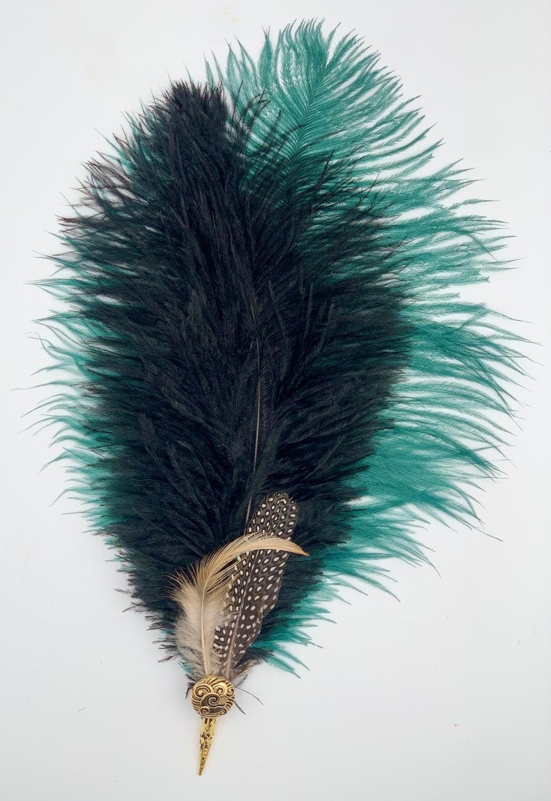 Op de afbeelding: Een zwarte en turquoise struisvogelveer met een gouden decoratief accent. De veer is lang en vloeiend, met een delicate textuur.
