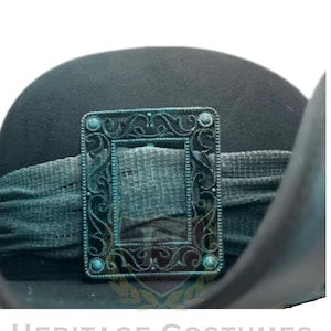 Puede incluir: Un sombrero negro con una banda de tela texturizada de color verde oscuro adornada con una hebilla rectangular de metal color cobre oxidado. La hebilla presenta intrincados patrones de volutas y peque&ntilde;os detalles circulares. El texto HERITAGE COSTUMES aparece debajo.