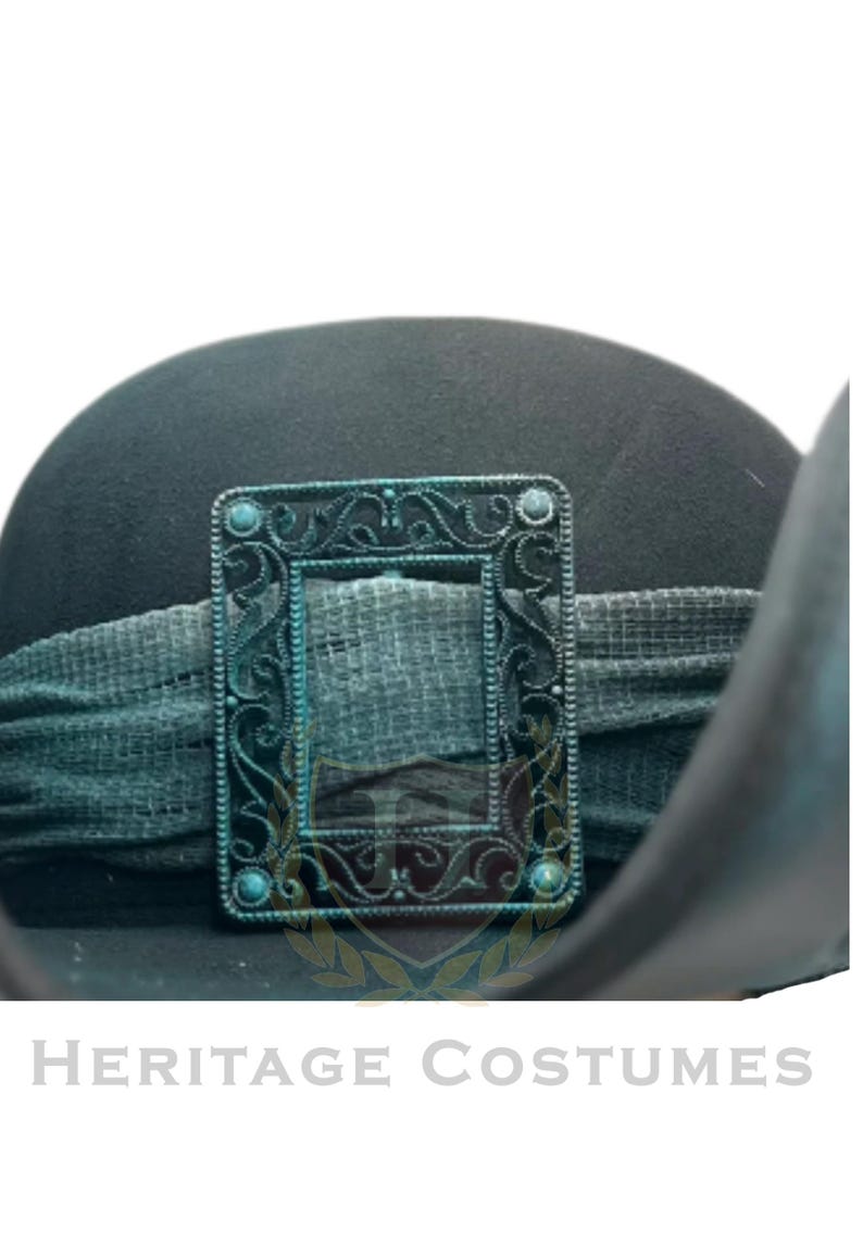 Puede incluir: Un sombrero negro con una banda de tela texturizada de color verde oscuro adornada con una hebilla rectangular de metal color cobre oxidado. La hebilla presenta intrincados patrones de volutas y peque&ntilde;os detalles circulares. El texto HERITAGE COSTUMES aparece debajo.