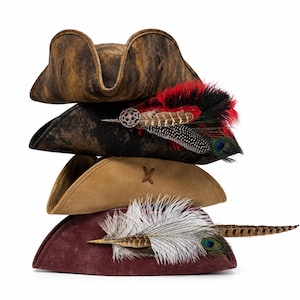 May include: A stack of four tricorn hats in various colors and textures. The top hat is distressed brown, the second is black with red and black feathers and a circular brooch, the third is tan, and the bottom is maroon with white and brown feathers.
