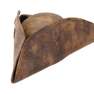 May include: A brown tricorn hat with a distressed, weathered texture. The hat features a rounded crown and a wide, upturned brim that is folded into a triangular shape. A small, stitched X-shaped detail is visible on the side of the brim.