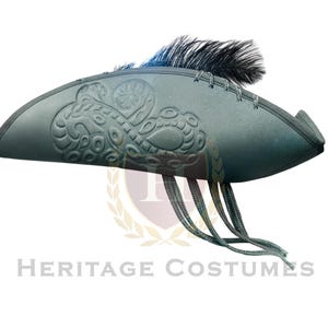 Puede incluir: Un sombrero de tres picos de color verde oscuro con un patr&oacute;n de remolinos en relieve en el lateral, una pluma negra en la parte superior y varios cordones verdes finos que cuelgan del ala. El sombrero tiene una forma estructurada y curva.