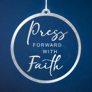 May include: A clear, circular ornament with a silver border and a white string for hanging. The ornament features the words "Press Forward With Faith" in elegant script against a dark blue background. Ideal for inspirational decor.