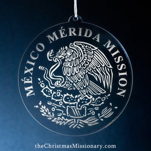 May include: Clear, circular Christmas ornament with the words "MÉXICO MÉRIDA MISSION" etched around the edge. The ornament features the Mexican coat of arms in the center. A white string is attached for hanging.