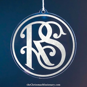 Könnte beinhalten: Klarer, kreisförmiger Weihnachtsschmuck mit den verschlungenen Buchstaben "RS" in einer dekorativen Schriftart. Der Schmuck wird an einer weißen Schnur vor einem dunkelblauen Hintergrund aufgehängt. Der Text "theChristmasMissionary.com" befindet sich unten.