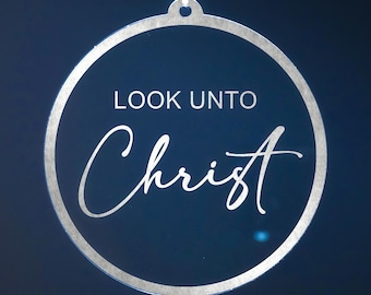 Puede incluir: Adorno circular con un borde metálico plateado y las palabras "LOOK UNTO Christ" en escritura blanca. El adorno cuelga de una cuerda blanca sobre un fondo azul oscuro.